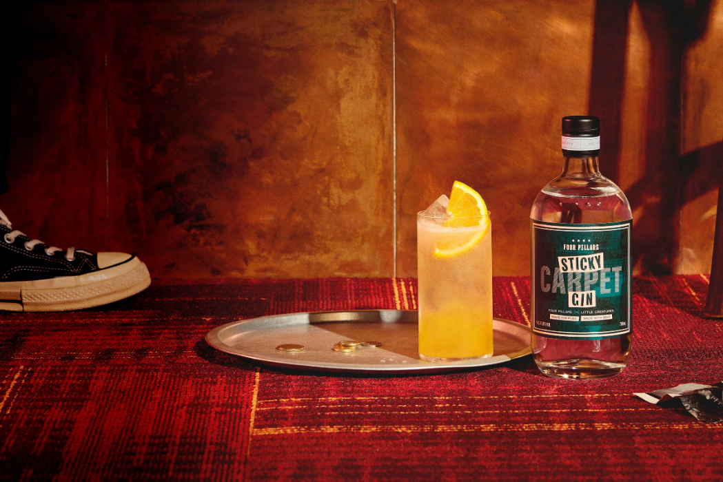 a bottle of Four Pillars Gin Sticky Carpet Gin on a red pub carpet floor next to a bar tray with a shakey pete gin cocktail and a converse sneaker. 
